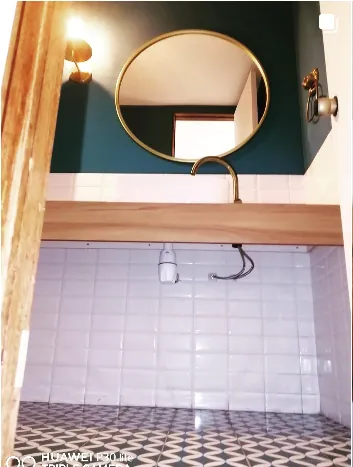 Baño con muro verde oscuro, espejo redondo dorado y cerámica blanca
